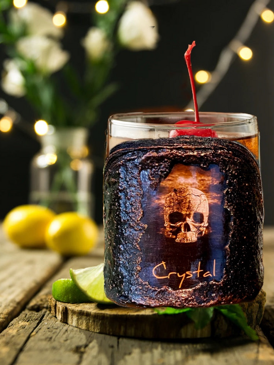 Glass with a dark liquid and skull design, garnished with a cherry, on a wooden surface with blurred lights in the background.
