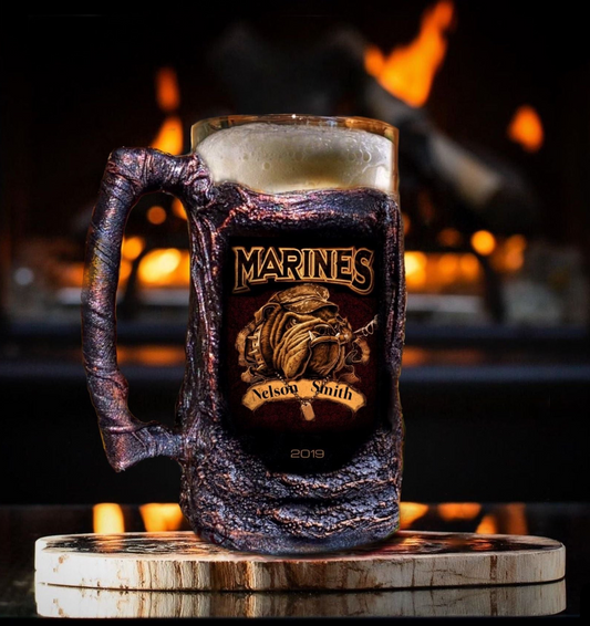 A beer mug with a personalized US Marines emblem and the text 'MARINES' on the front, placed on a wooden slice with a fire in the background.